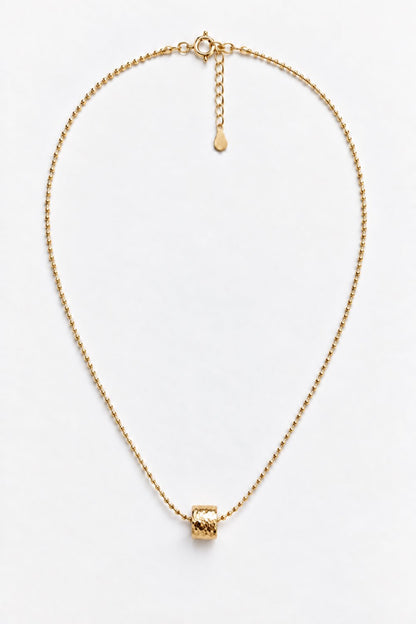 A delicate gold chain necklace with a small, textured cylindrical bead pendant, displayed on a clean white background.