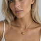 A close-up of a blonde woman wearing a delicate gold chain necklace with a small textured cube pendant, paired with a cream ribbed tank top against a neutral gray background.