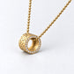 A gold pendant necklace featuring a textured cylindrical ring with a border of small clear stones, suspended from a gold ball chain on a light gray background.