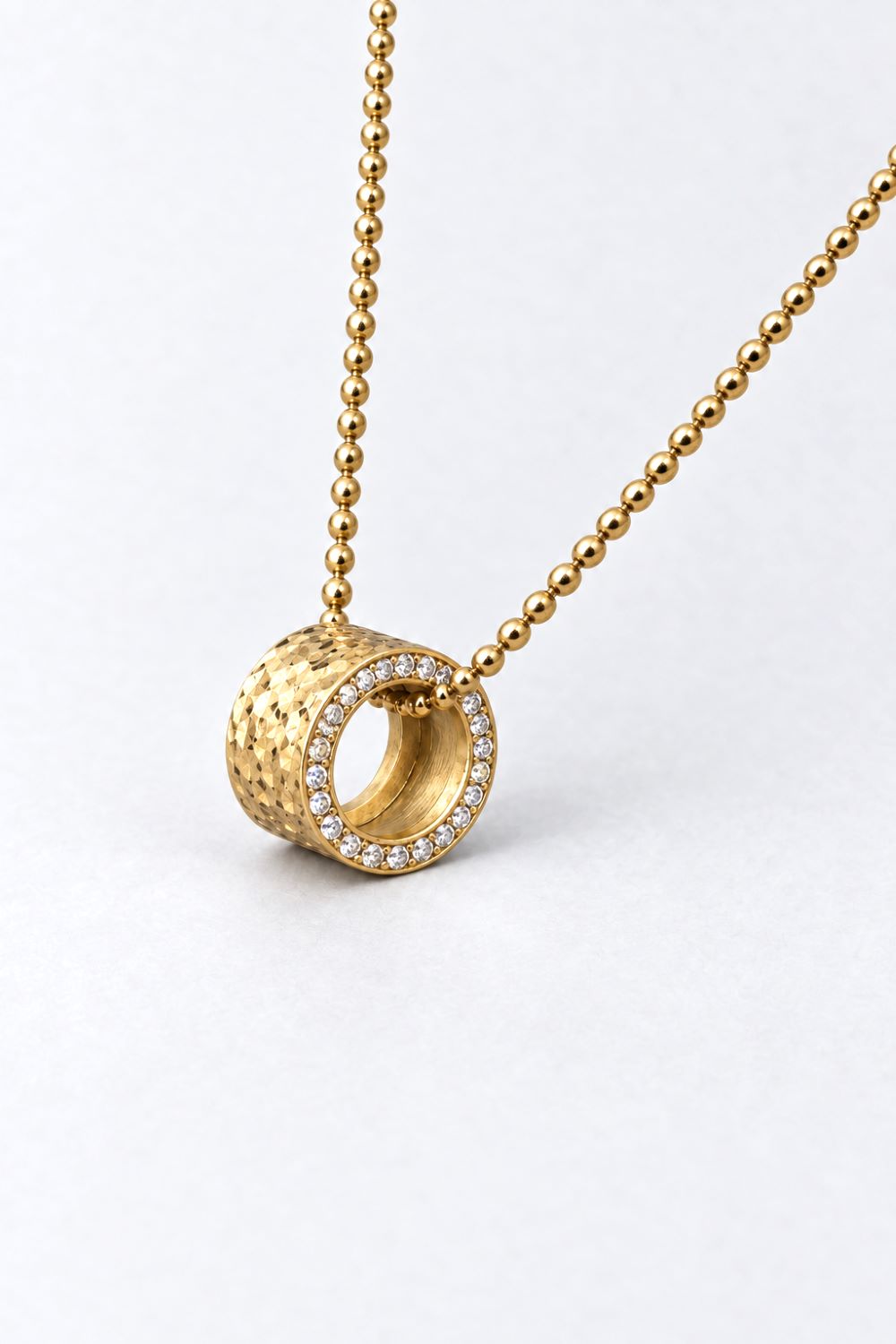 A gold pendant necklace featuring a textured cylindrical ring with a border of small clear stones, suspended from a gold ball chain on a light gray background.