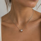 A close-up of a model wearing a delicate silver chain necklace with a small cylindrical silver pendant featuring a clear, sparkling center stone.