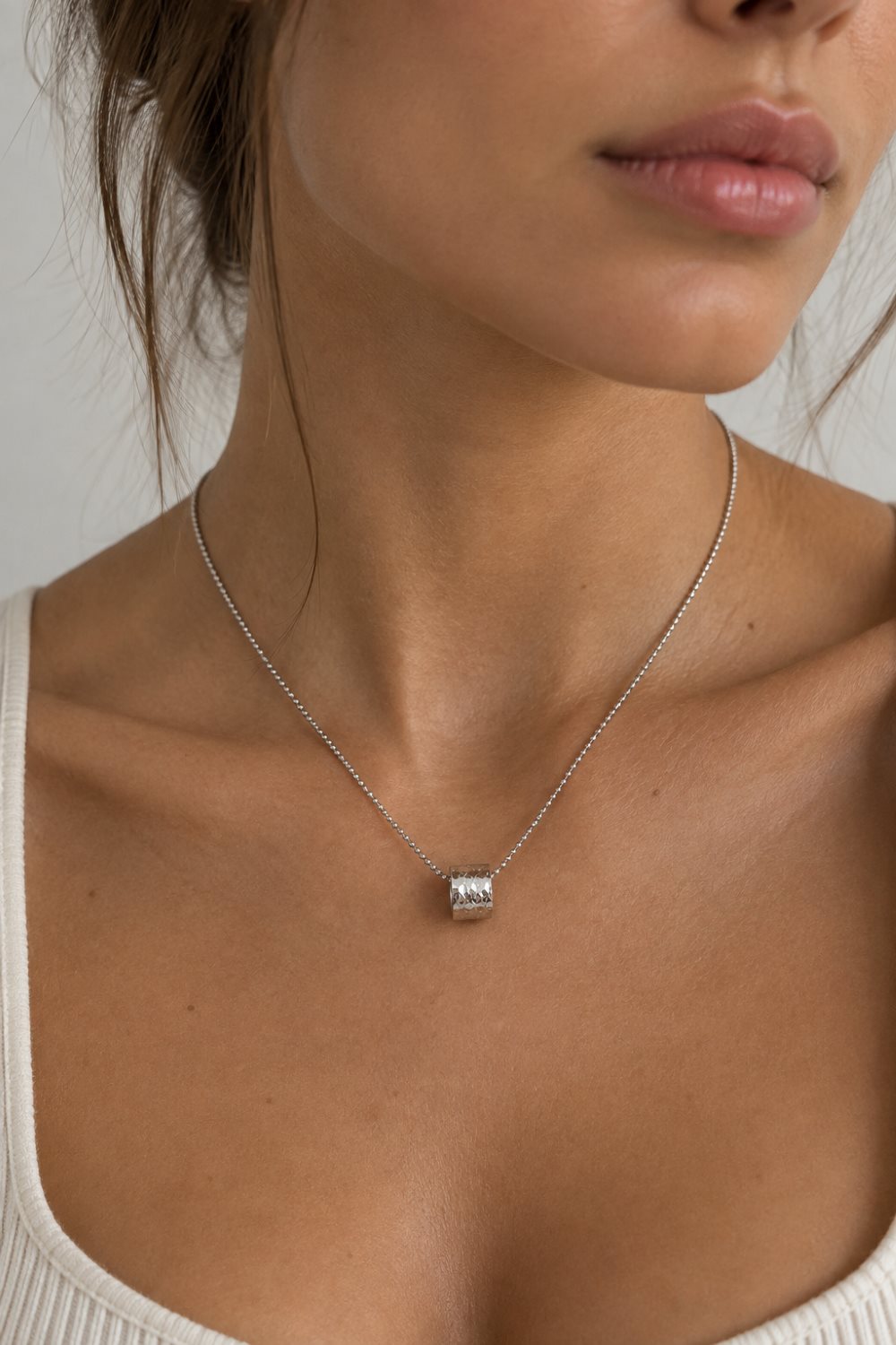 A close-up of a model wearing a delicate silver chain necklace with a small cylindrical silver pendant featuring a clear, sparkling center stone.
