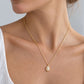 A close-up of a woman wearing a delicate gold chain necklace with a small oval clear stone pendant, paired with a white top against a light gray background.