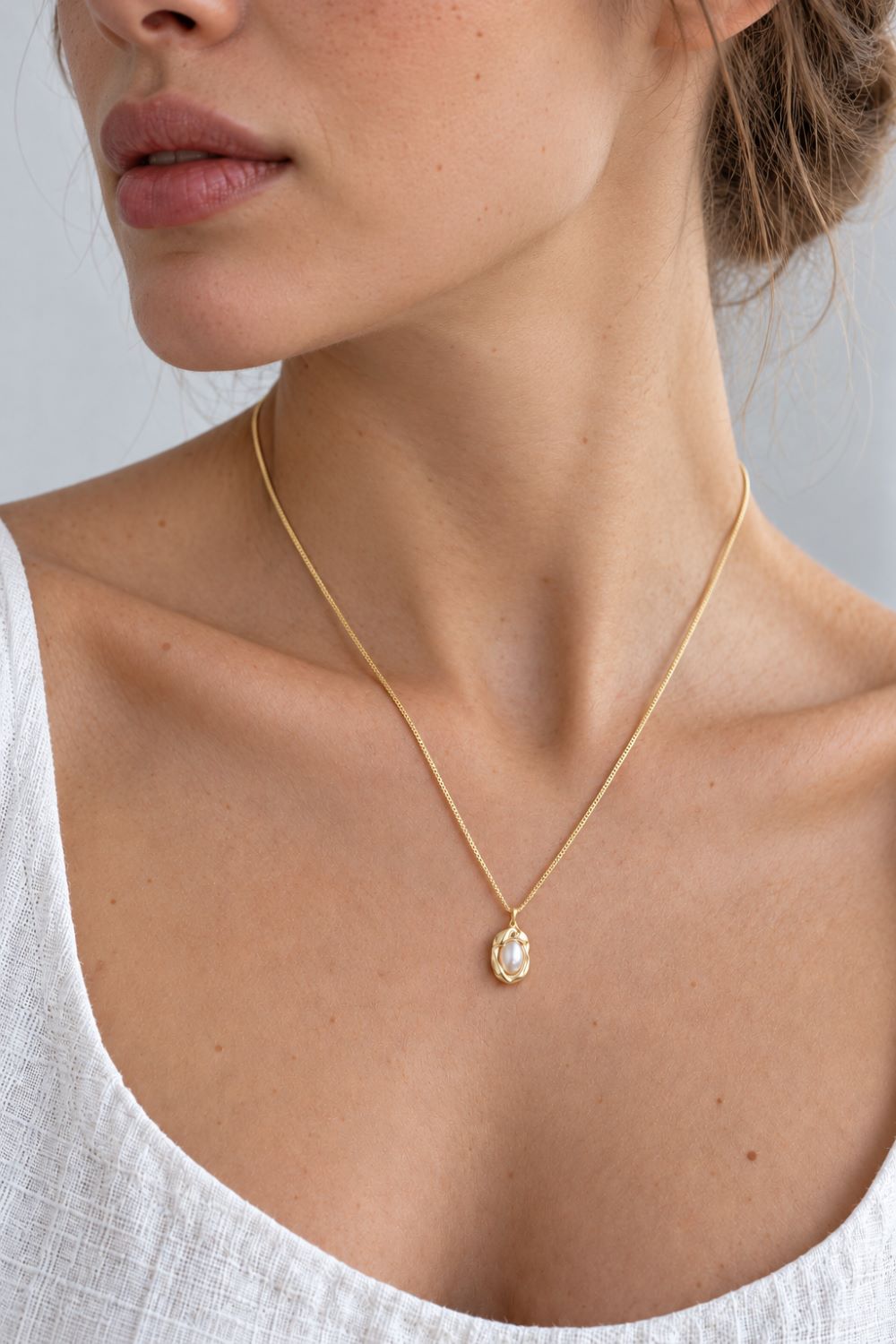 A close-up of a woman wearing a delicate gold chain necklace with a small oval clear stone pendant, paired with a white top against a light gray background.
