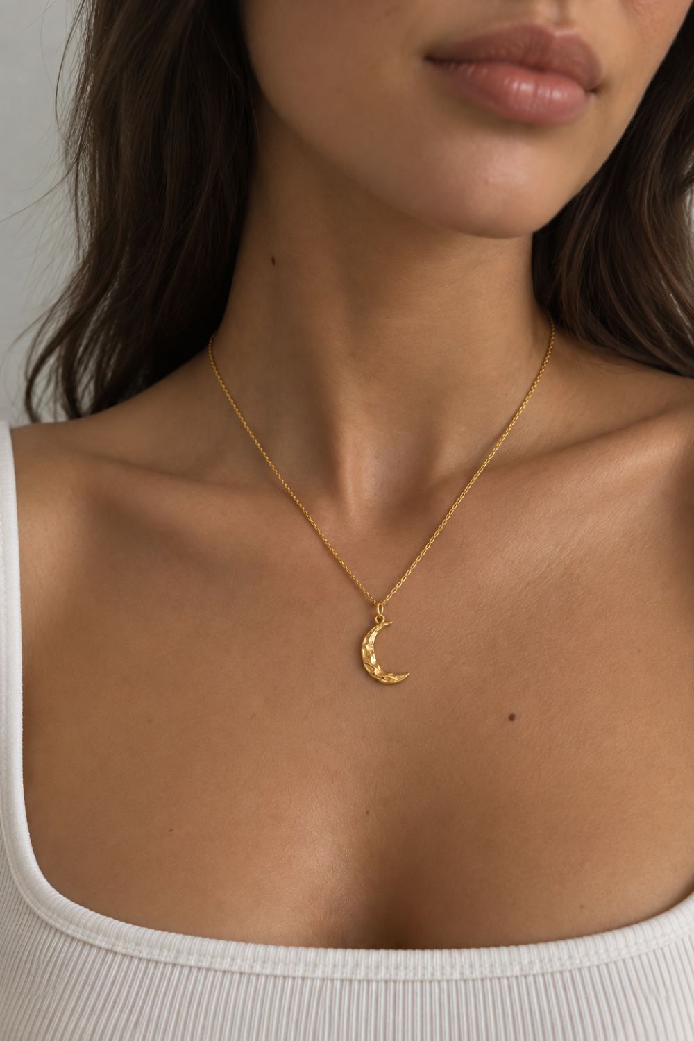 A close-up of a model wearing a delicate gold chain necklace with a crescent moon pendant, styled with a white ribbed tank top.
