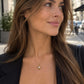 A woman with long brown hair wearing a delicate gold teardrop pendant necklace, styled with a black top and blazer, photographed outdoors on a city street.