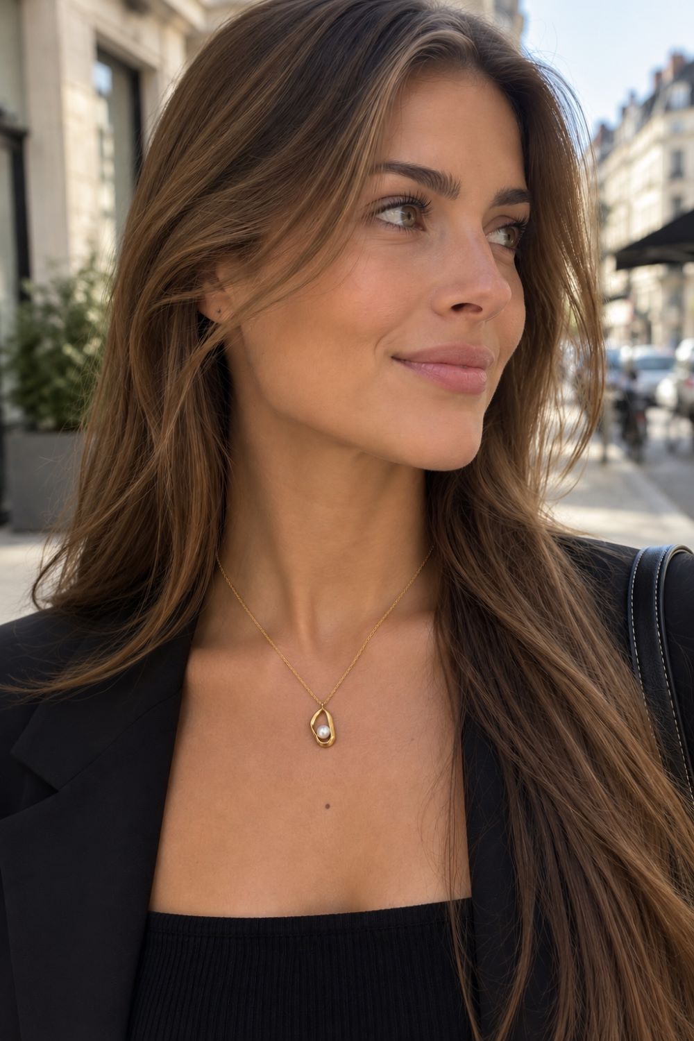 A woman with long brown hair wearing a delicate gold teardrop pendant necklace, styled with a black top and blazer, photographed outdoors on a city street.