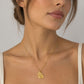 A close-up of a woman wearing a delicate gold chain necklace with a small, textured gold pendant, paired with a cream-colored ribbed tank top against a neutral gray background.