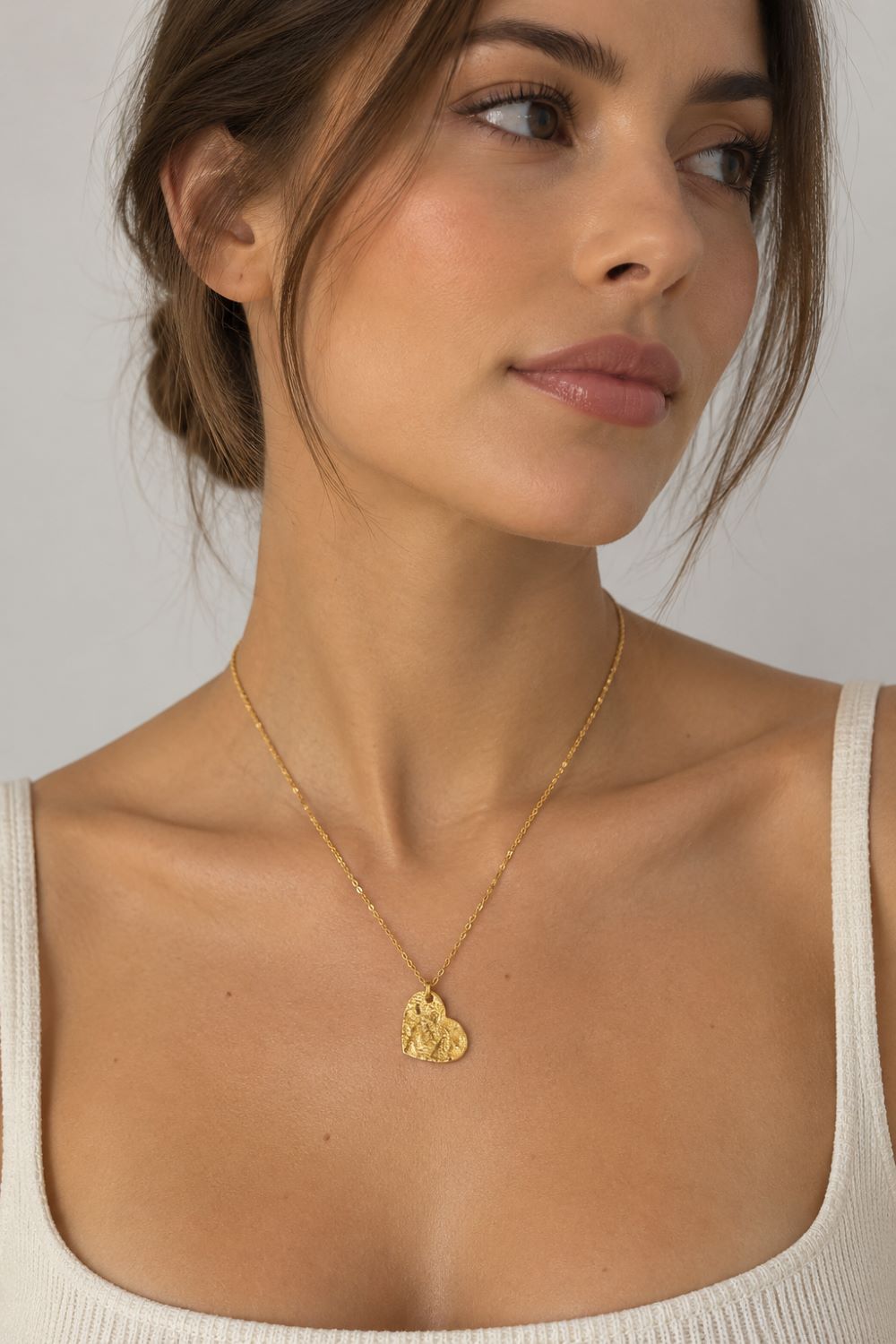 A close-up of a woman wearing a delicate gold chain necklace with a small, textured gold pendant, paired with a cream-colored ribbed tank top against a neutral gray background.