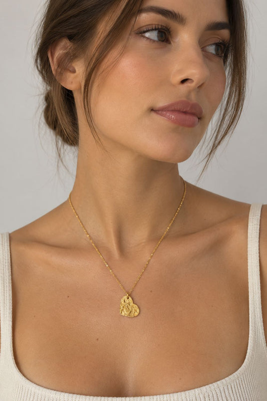 A close-up of a woman wearing a delicate gold chain necklace with a small, textured gold pendant, paired with a cream-colored ribbed tank top against a neutral gray background.