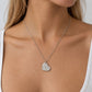 A close-up of a woman wearing a delicate silver chain necklace with a textured, hammered heart-shaped pendant. She is wearing a white ribbed tank top against a neutral light gray background.