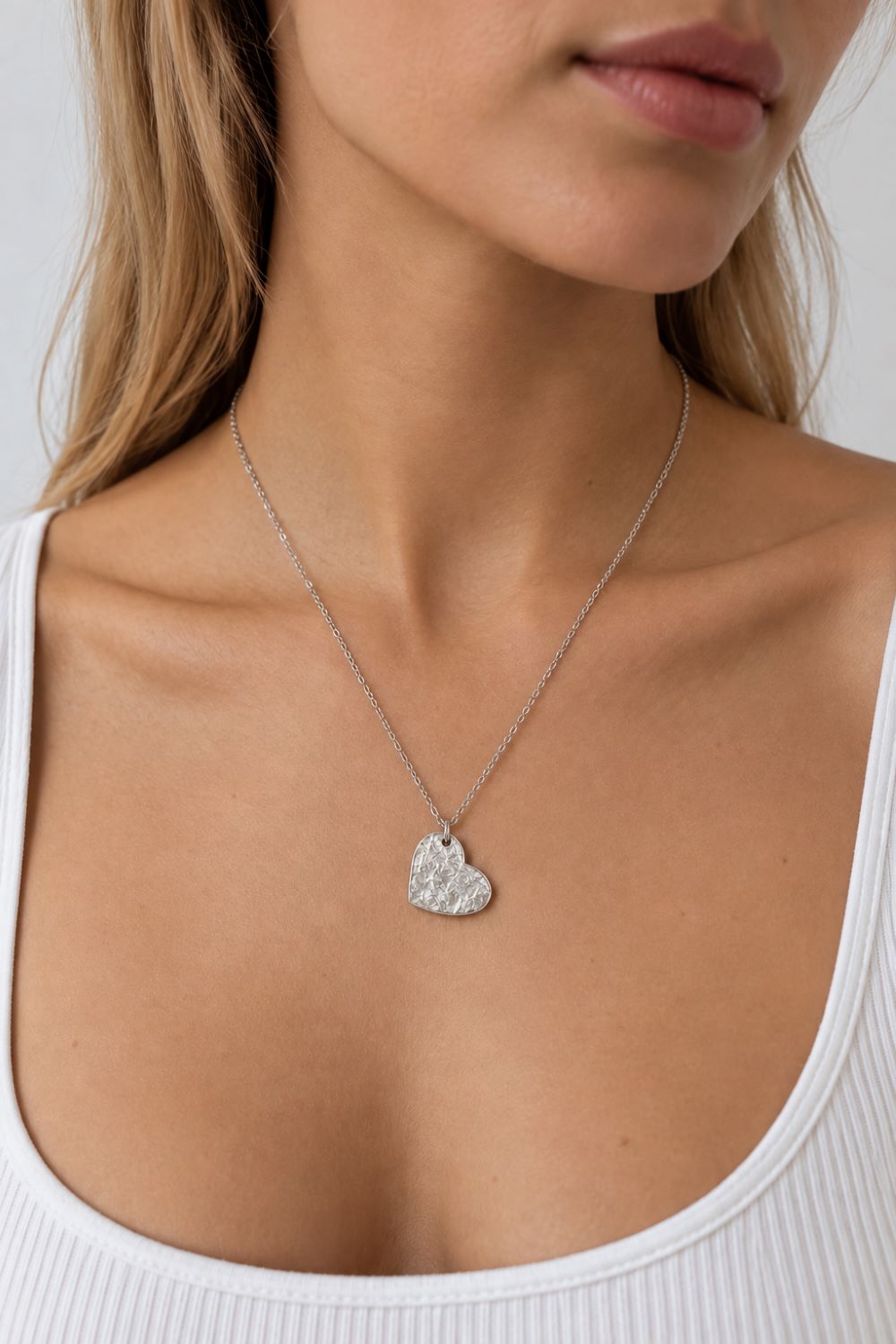 A close-up of a woman wearing a delicate silver chain necklace with a textured, hammered heart-shaped pendant. She is wearing a white ribbed tank top against a neutral light gray background.