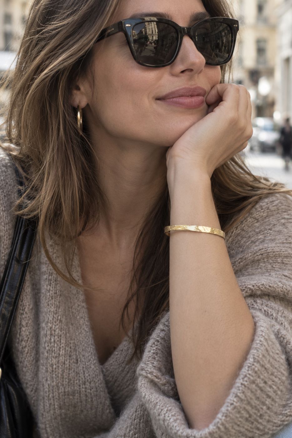 A woman wearing black cat-eye sunglasses, a gold hoop earring, and a gold bangle bracelet, posing outdoors in a beige knit sweater.