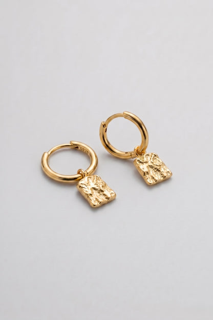 A pair of gold huggie hoop earrings with textured, irregularly shaped dangling charms, displayed on a clean light gray background.