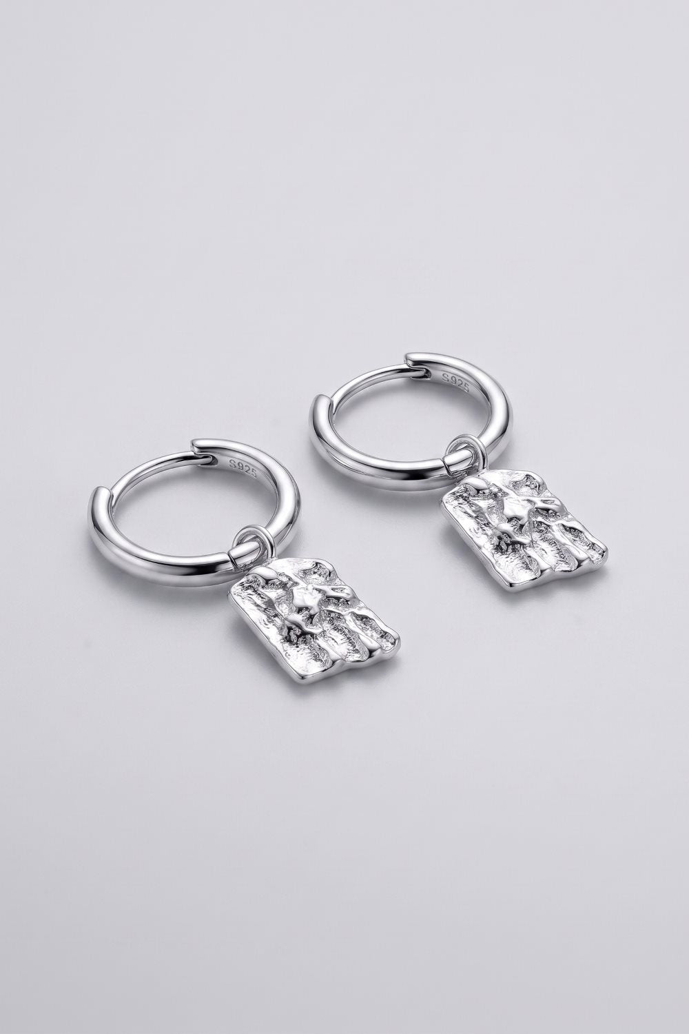 A pair of silver huggie hoop earrings with textured rectangular dangling charms, displayed on a clean light gray background.