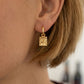 A close-up of a person's ear wearing a small gold drop earring with a textured square pendant.