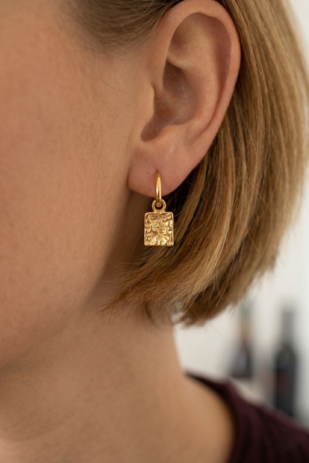 A close-up of a person's ear wearing a small gold drop earring with a textured square pendant.