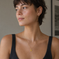 A woman with short dark hair wearing a delicate gold necklace with a small gold pendant, styled with a black ribbed tank top in a softly lit indoor setting.