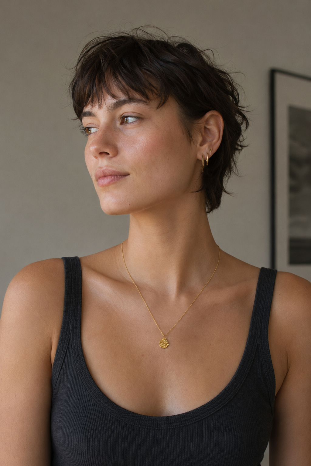 A woman with short dark hair wearing a delicate gold necklace with a small gold pendant, styled with a black ribbed tank top in a softly lit indoor setting.
