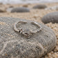 A silver-toned open ring with a central knot-like charm and textured ends, resting on a large smooth beach stone with sand, rocks, and the ocean blurred in the background.