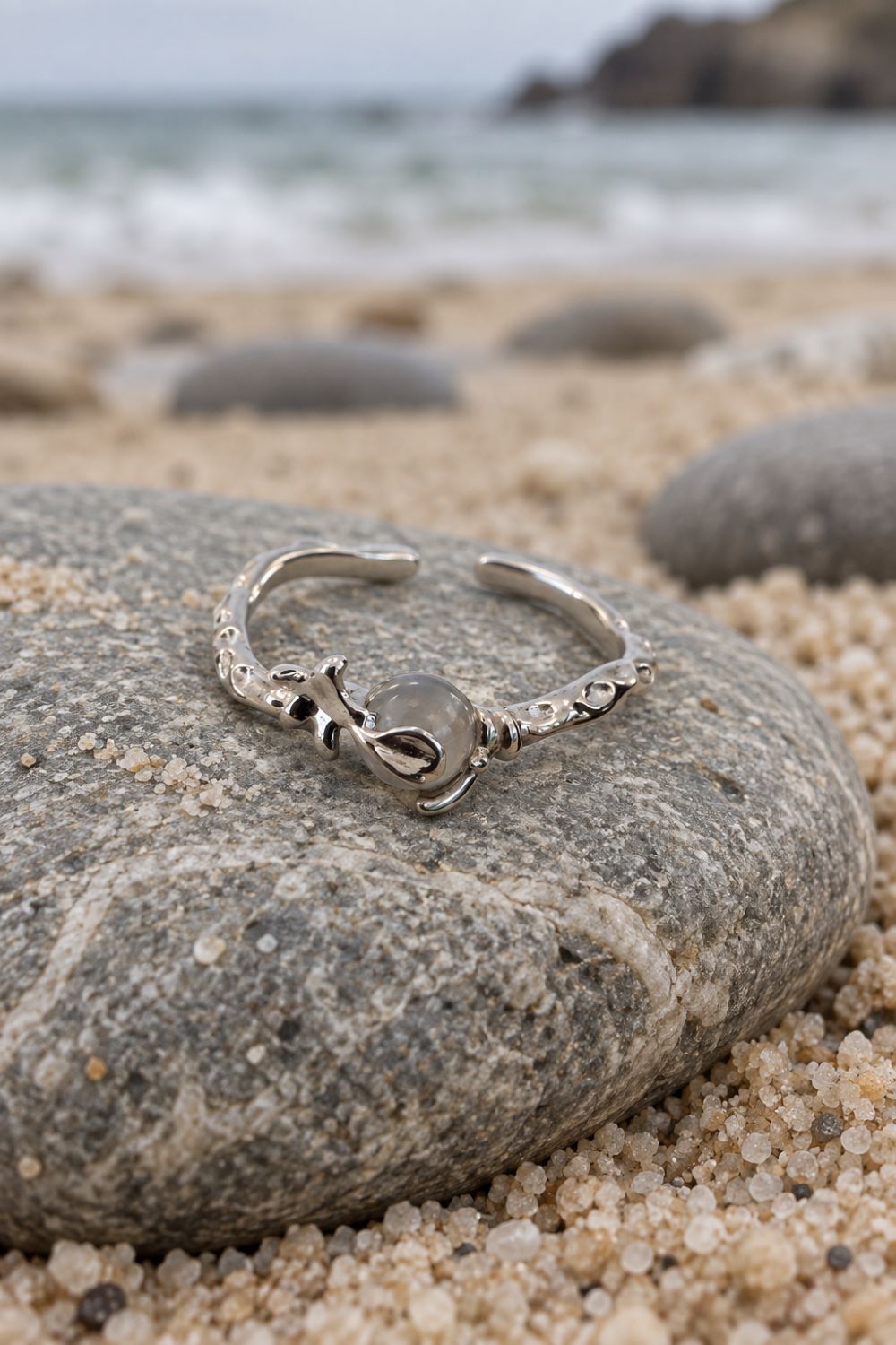 A silver-toned open ring with a central knot-like charm and textured ends, resting on a large smooth beach stone with sand, rocks, and the ocean blurred in the background.