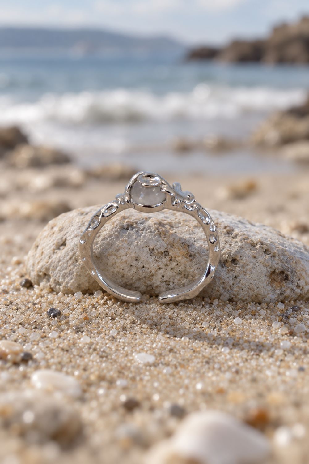 A silver open ring with a clear teardrop-shaped gemstone and small clear stones along the band, resting on a sandy beach with a blurred ocean background.
