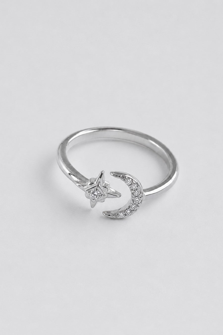 A silver-toned ring featuring a crescent moon and star design, both adorned with small clear stones, set against a plain light gray background.