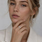 A close-up portrait of a woman wearing a silver crescent moon ring on her finger, dressed in a white blazer against a soft light gray background.