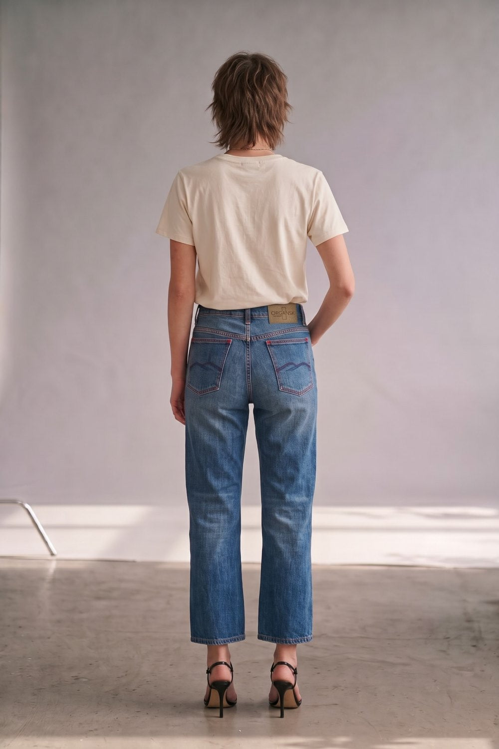 A model viewed from behind wearing blue cropped jeans, a cream t-shirt, and black high-heeled sandals in a minimalist studio setting.
