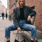A woman sits on a bench in an outdoor urban setting, wearing an oversized dark gray blazer, black top, blue cropped jeans, black ankle boots, and holding a black handbag.
