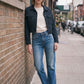 A woman stands on a city sidewalk wearing a dark denim jacket, a cream-colored top, and blue jeans, posing next to a brick wall.