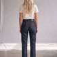 A woman viewed from behind wearing dark indigo cropped jeans with red contrast stitching, a cream t-shirt, and black sandals in a minimalist studio setting.