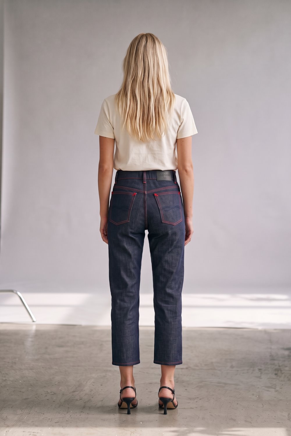 A woman viewed from behind wearing dark indigo cropped jeans with red contrast stitching, a cream t-shirt, and black sandals in a minimalist studio setting.