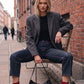 A woman sits on a bench in an outdoor urban setting, wearing an oversized dark gray blazer, black top, dark blue jeans, and red pointed-toe ankle boots, with a black handbag resting in front of her.