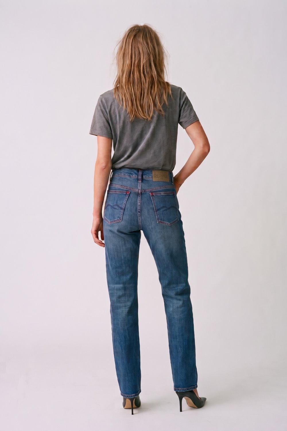 A woman viewed from behind wearing blue denim jeans, a dark gray t-shirt, and black high heels against a light gray studio background.