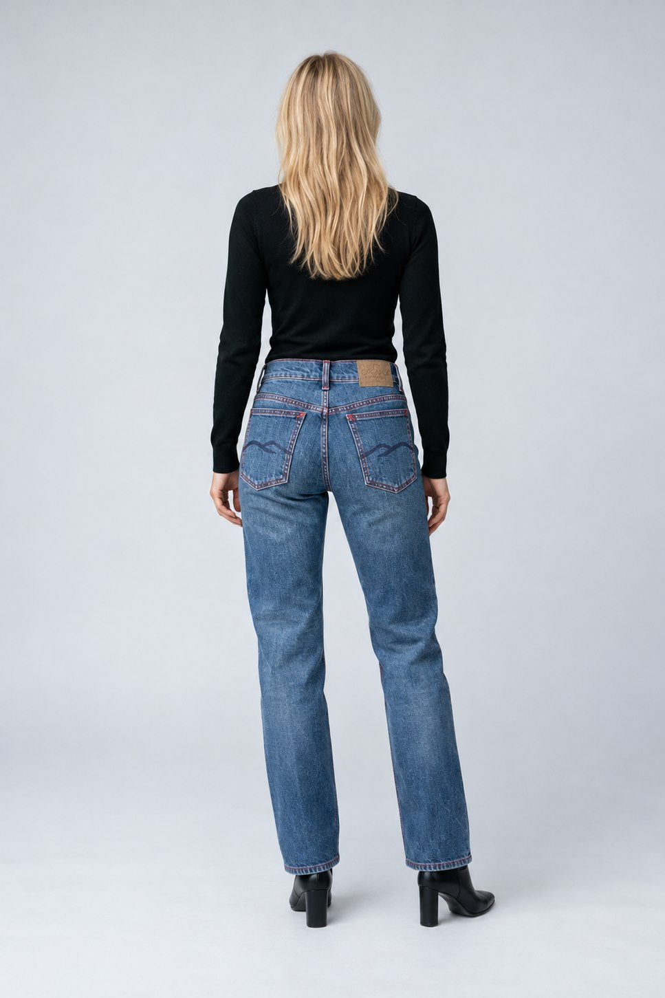 A woman viewed from behind wearing blue straight-leg jeans, a black long-sleeve top, and black heeled boots against a light gray studio background.