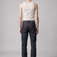 A man viewed from behind wearing dark indigo denim jeans with red stitching, a cream-colored ribbed tank top, and black shoes, standing against a light gray studio background.