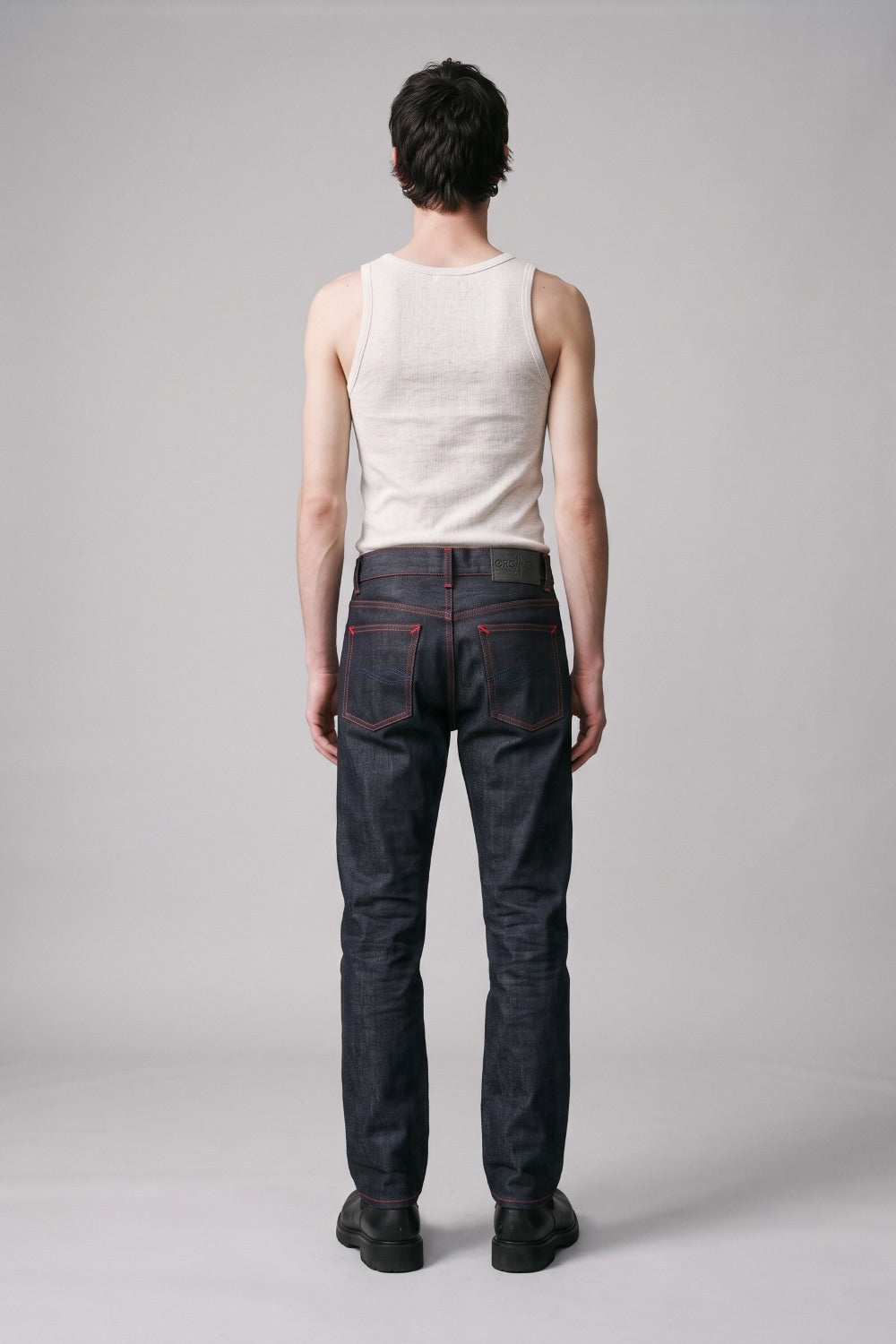 A man viewed from behind wearing dark indigo denim jeans with red stitching, a cream-colored ribbed tank top, and black shoes, standing against a light gray studio background.