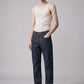 A male model stands against a light gray studio background wearing a fitted off-white tank top, dark blue straight-leg jeans, and black boots.