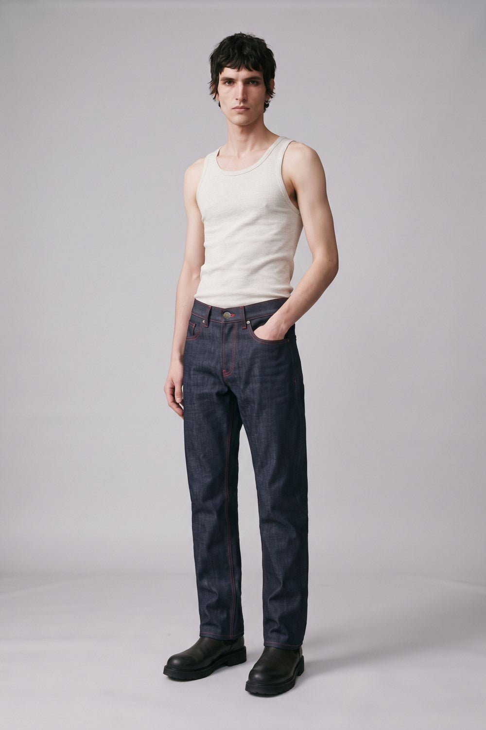 A male model stands against a light gray studio background wearing a fitted off-white tank top, dark blue straight-leg jeans, and black boots.