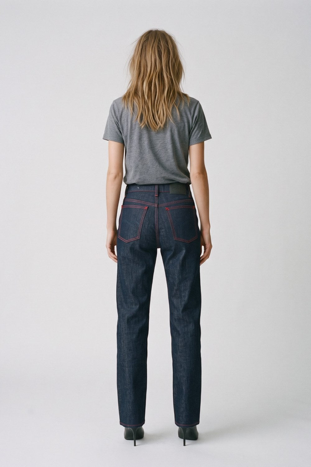 A woman viewed from the back wearing dark blue straight-leg denim jeans with red contrast stitching, a heather gray t-shirt, and black heels, against a light gray studio background.