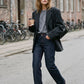 A woman walking on an urban street wearing a black blazer, dark gray t-shirt, dark blue straight-leg jeans, and black pointed-toe heels, holding a coffee cup.