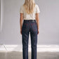 A woman viewed from behind wearing dark blue straight-leg jeans with red contrast stitching, a cream t-shirt, and black heels, standing in a minimalist studio setting.