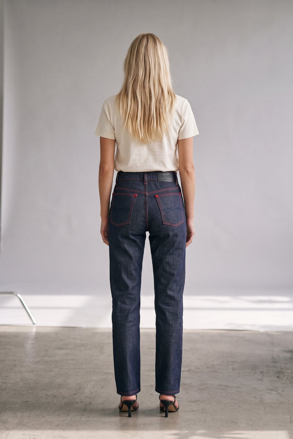 A woman viewed from behind wearing dark blue straight-leg jeans with red contrast stitching, a cream t-shirt, and black heels, standing in a minimalist studio setting.
